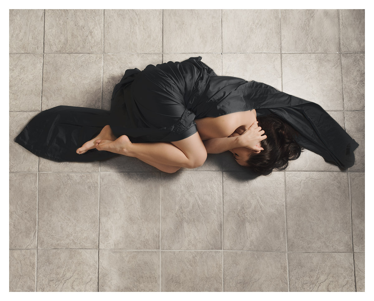 A woman wrapped in a black sheet lies on a tile floor. 
