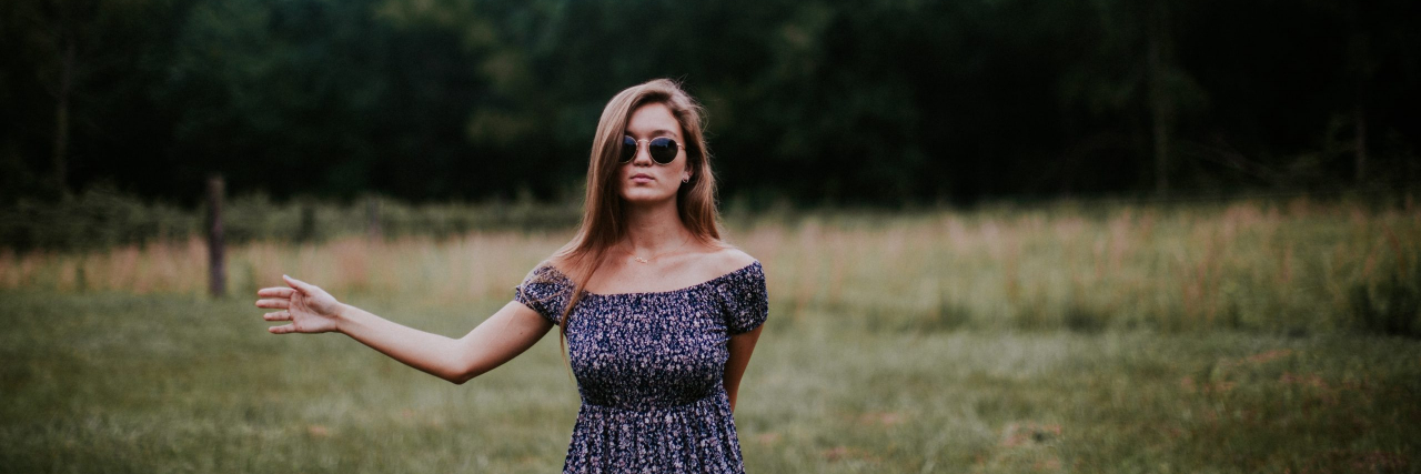 The Truth About Living Openly With a Mental Illness A woman with brown hair and sunglasses wearing a black and white dress extending her arm to the side, standing in front of a field and forest.