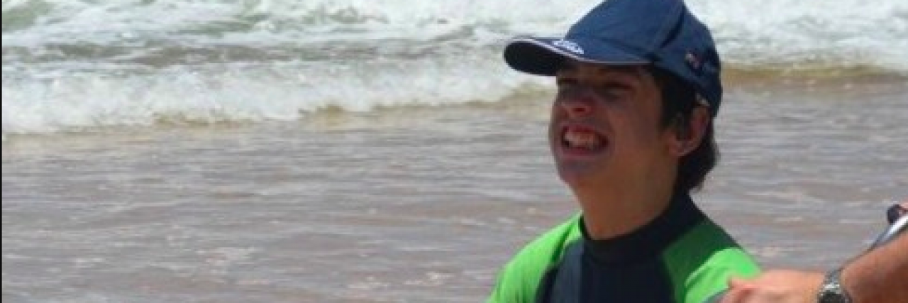 A boy in a wheelchair, wearing a hat, with someone helping push his wheelchair by the ocean