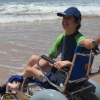 A boy in a wheelchair, wearing a hat, with someone helping push his chair by the ocean