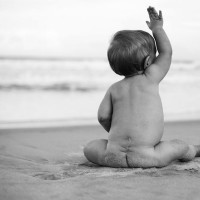 Black and white photo of baby sitting on the beach with arm raised