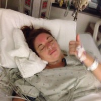 Kerri lying in a hospital bed with her eyes closed, giving the thumbs up