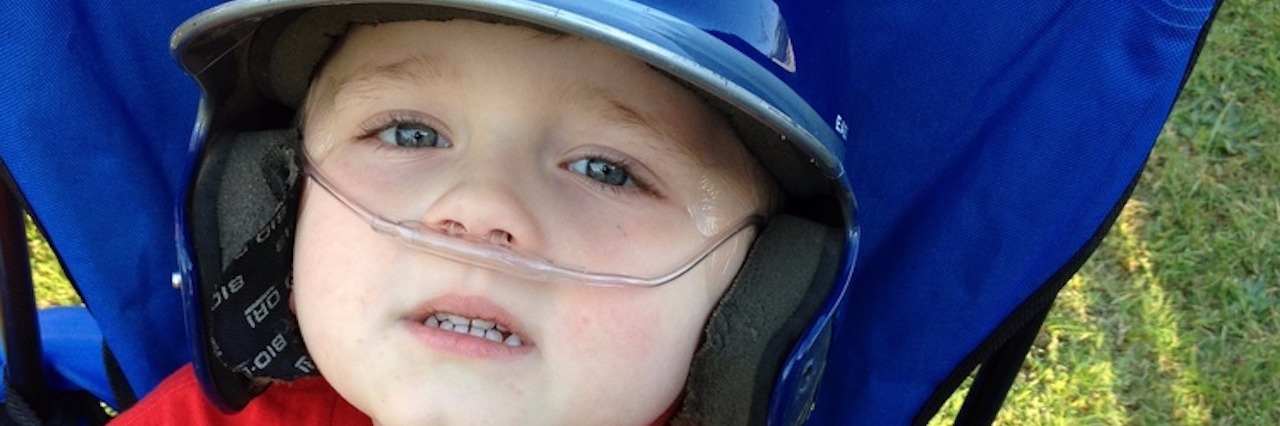 Playing T-Ball With Mitochondrial Disease young boy sitting in chair outside and wearing baseball helmet