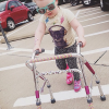 A small girl wearing sunglasses uses a walker to cross a parking lot
