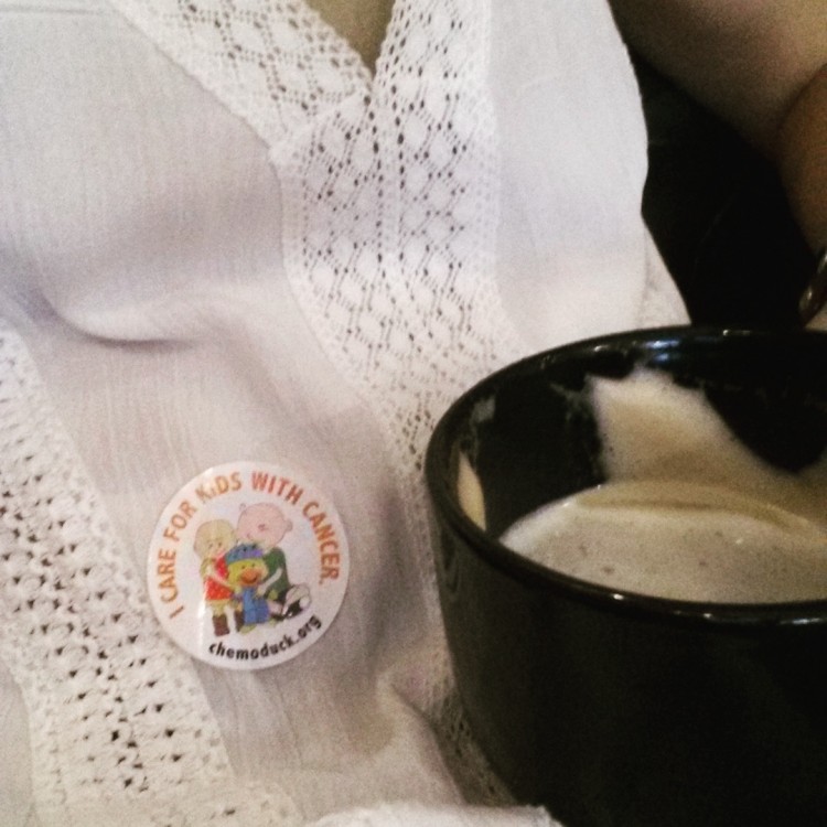 A close up of a woman holding a cup of coffee. On her shirt is a sticker that reads "I care for kids with cancer."