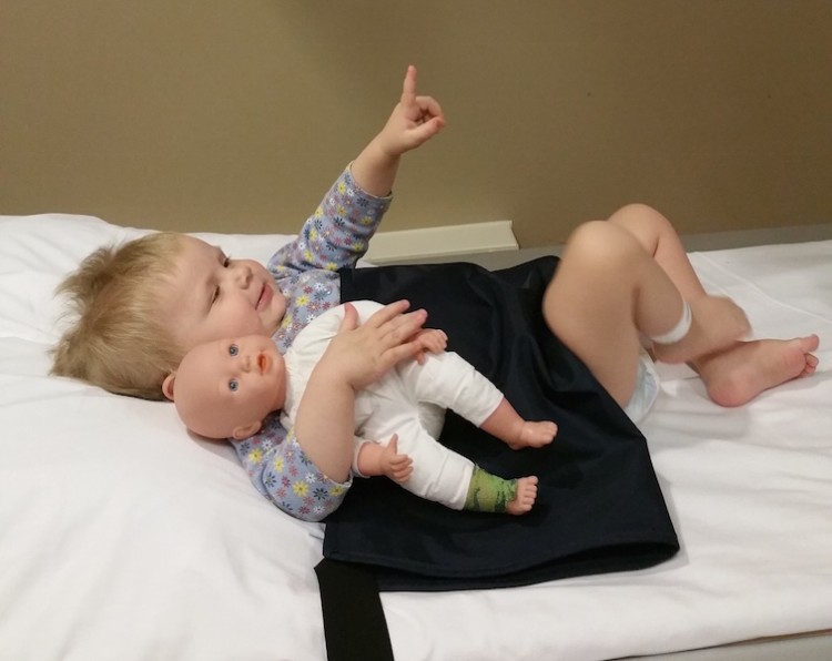 A little girl laying on a hospital bed, holding a little doll and pointing upward.