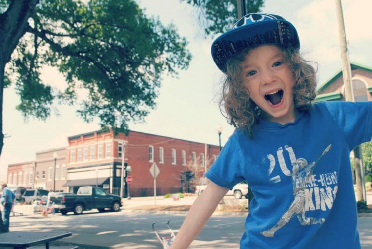 Young boy outside with a hat and long, curly hair. His mouth is wide open in a smile.