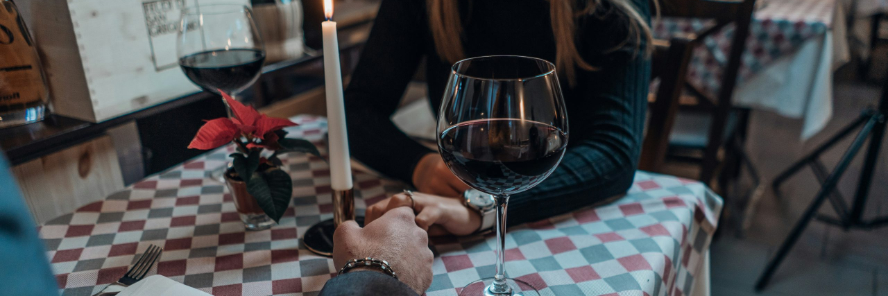 A romantic date for two with a couple holding hands across a checkered red, white, and gray table with a plant, wine glasses, a candle, and phone on top.