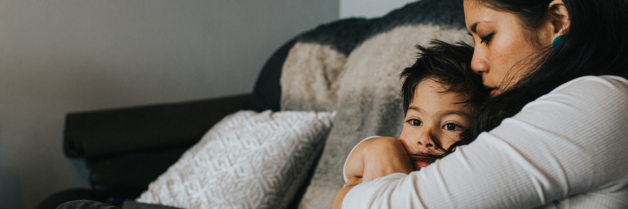 How to Help Kids Cope With Anxiety A mother and son sit on a sofa in a house environment. She tenderly embraces him and kisses his head as he snuggles into her.