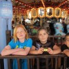 Three kids leaning over a rail at a merry-go-round