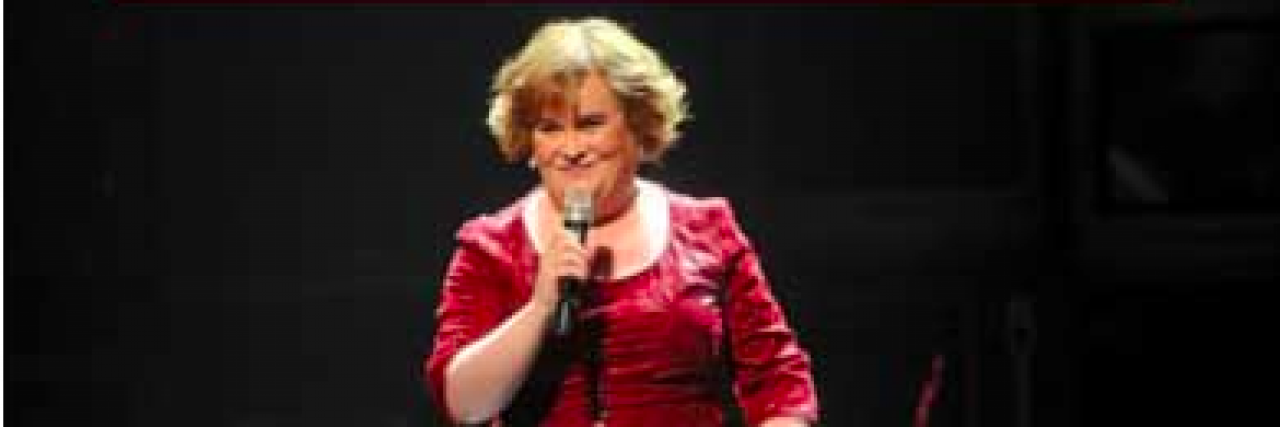 Susan Boyle standing on a darkly lit stage with a microphone