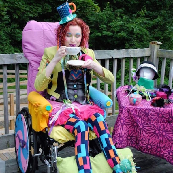 A young woman in a costume sipping on tea in her wheelchair next to a table covered with more costume iterms