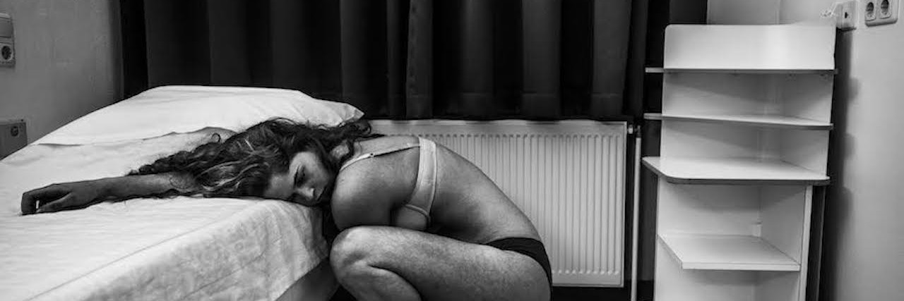 Woman crouching over with her head on the bed of her psychiatric hospital hotel room