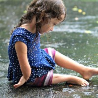 A small toddler sits in the rain looking at an object under her foot