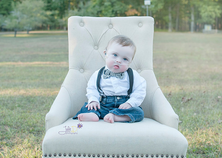 Baby wearing bow tie and suspenders sitting on a chair in a park