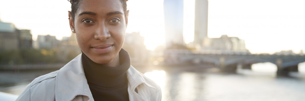 Black woman wearing a turtleneck and light jacket outside on a sunny day. She is standing beside a body of water with a cityscape in the background.
