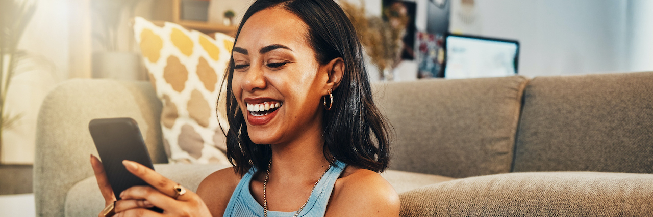 18 Memes That Nail What It's Like to Live With Chronic Illness Woman of color sitting on floor against a couch, holding her phone and laughing