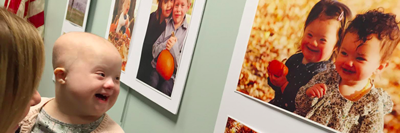 Young girl without hair smiles while she is held up to look at portrait of herself and friend on gallery wall