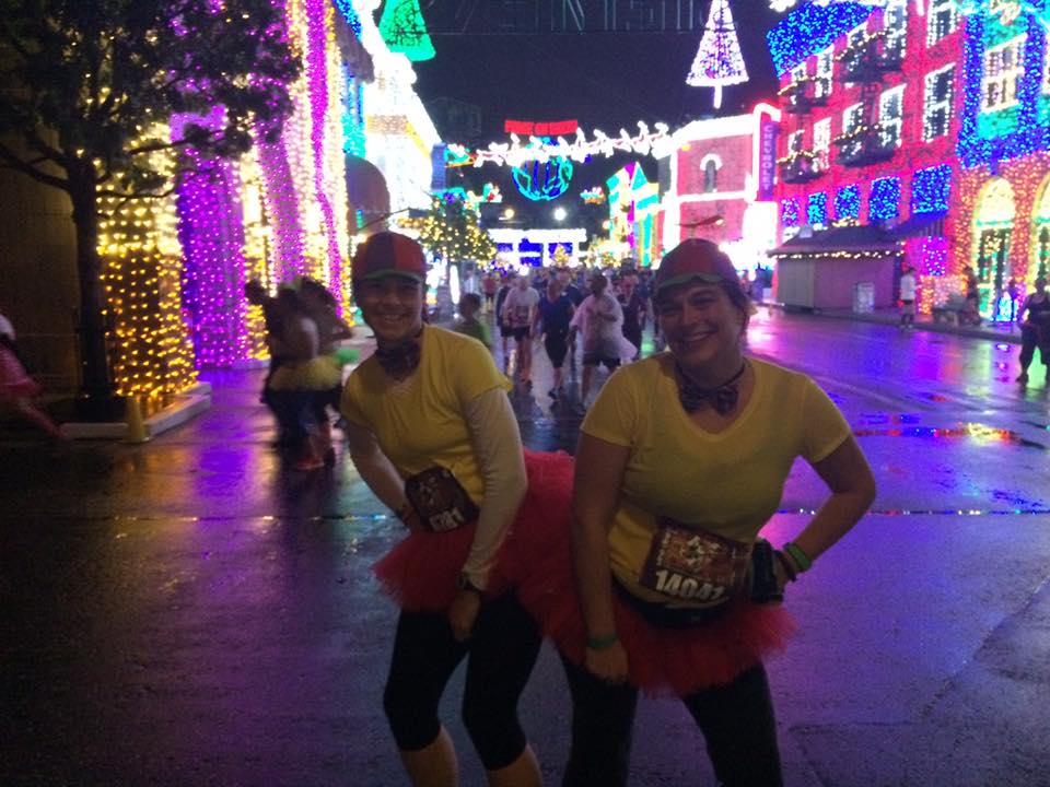 Two people dressed up on a street with lots of Christmas lights. 