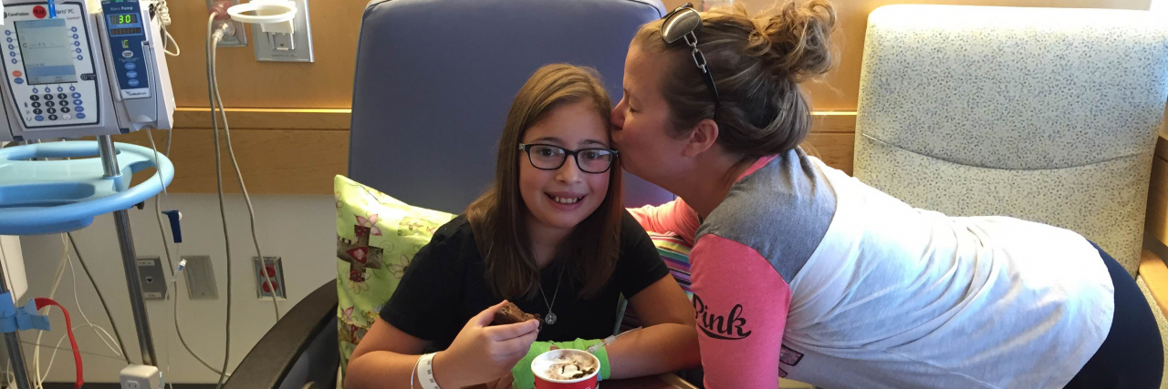 Mother kissing daughter on cheek while daughter sits in hospital chair