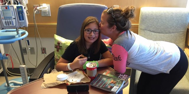 Mother kissing daughter on cheek while daughter sits in hospital chair