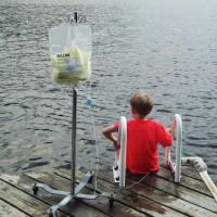A boy with medical equipment nearby sits with feet hanging over a body of water at a pier