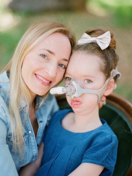 A mother smiles with her daughter, who's wearing a breathing tube. 