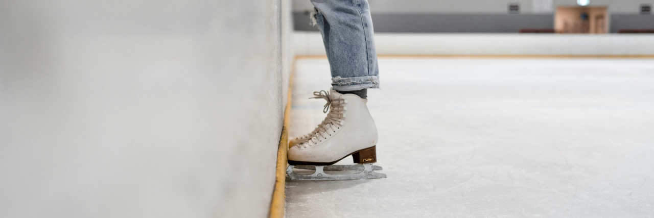 A person wearing jeans and white ice skates, standing on the inside of an iceskating ring against the wall.