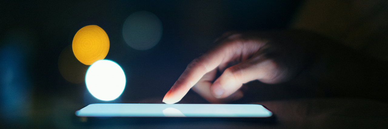 Texts to Send People With Anxiety Close-up shot of finger scrolling on smartphone screen in a dark environment, with spots of light in the background.