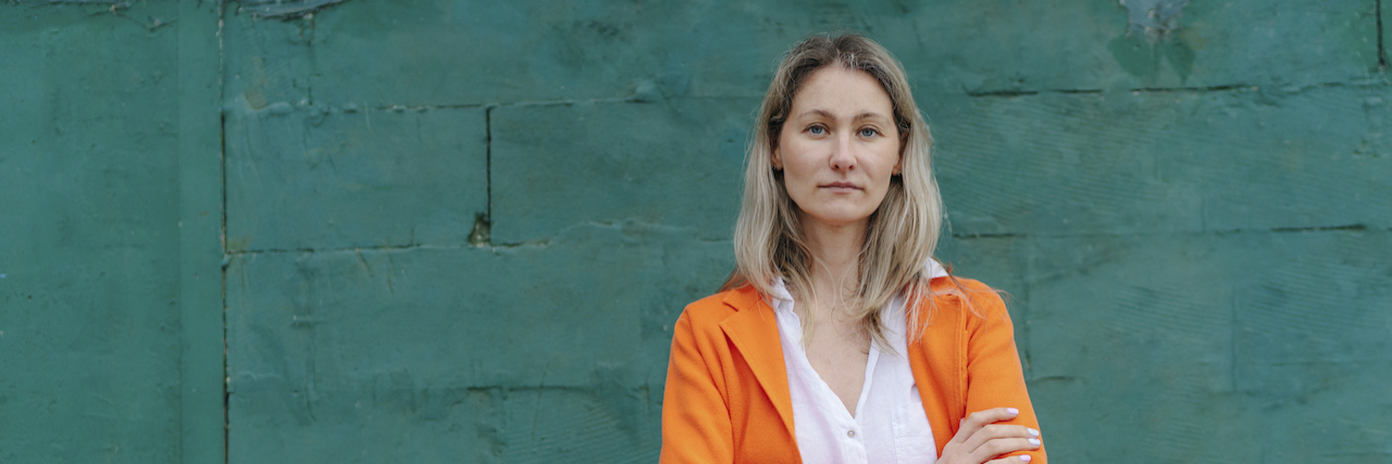 7 Stages I Experienced After My POTS Diagnosis Woman standing with arms crossed in front of green wall