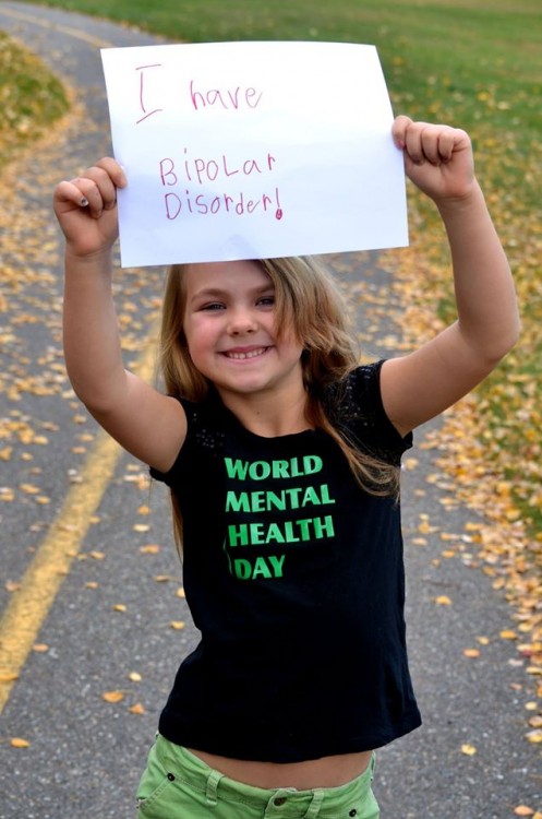 Jade's daughter is smiling and holding a sign that says, "I have bipolar disorder!" She has blonde hair, and is wearing a black shirt that says "World Mental Health Day" in green.