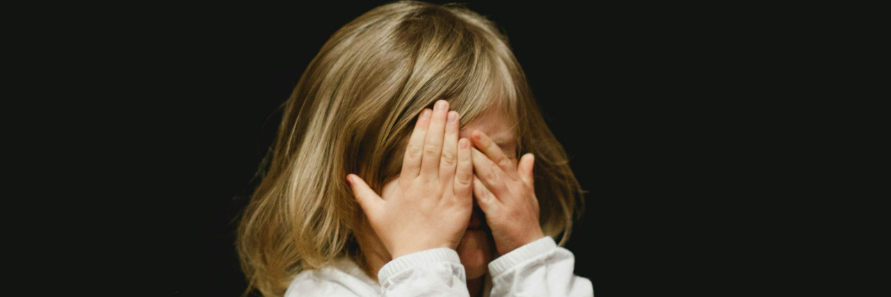 A young girl with dirty blonde hair and a white long sleeve shirt covering her face with her hands