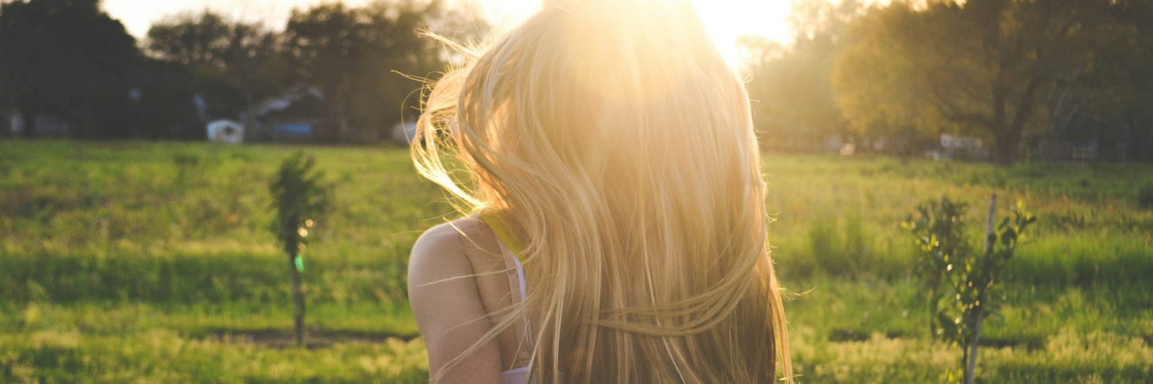 To the Little Girl Who Just Got Diagnosed With Bipolar Disorder A woman with blonde and brunette hair wearing a green top and black shorts with white flowers running away towards the sun across a field.