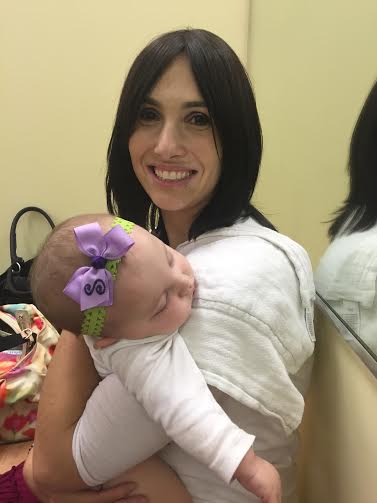 Randi smiling and holding her baby. Her baby is wearing a green headband with a purple bow.