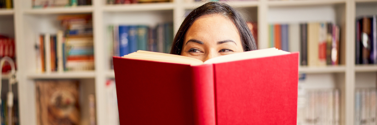 Woman's face peeking from behind an open book with full bookshelf in background