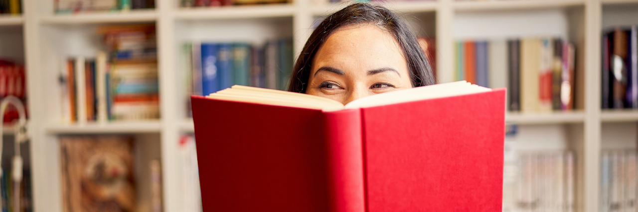 Book Recommendations for People With Chronic Illness Woman's face peeking from behind an open book with full bookshelf in background