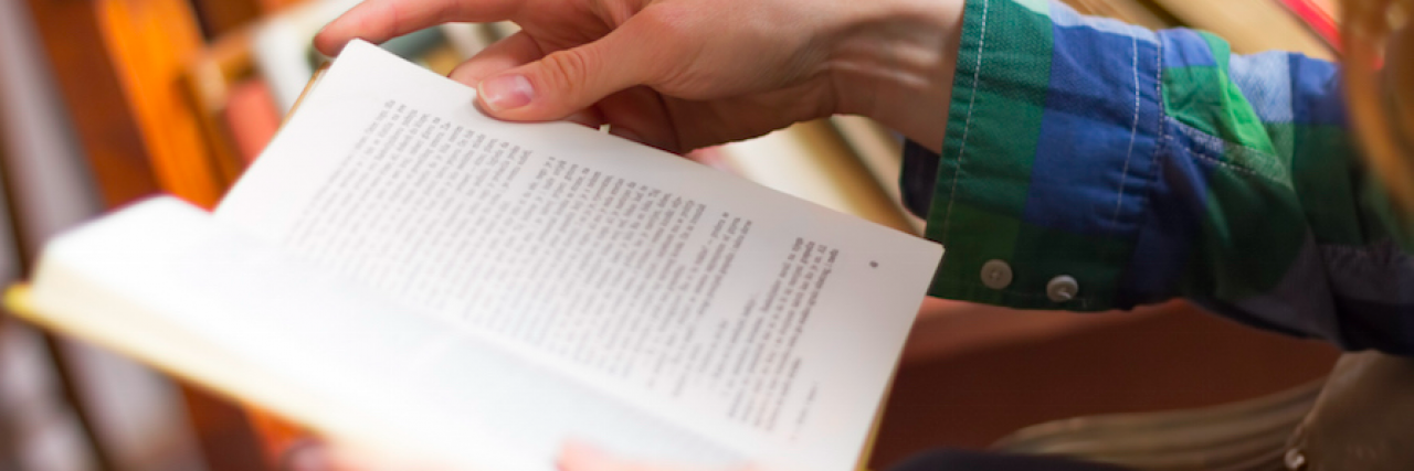 Close up of a person's hands holding an open book, with books on a shelf in the background