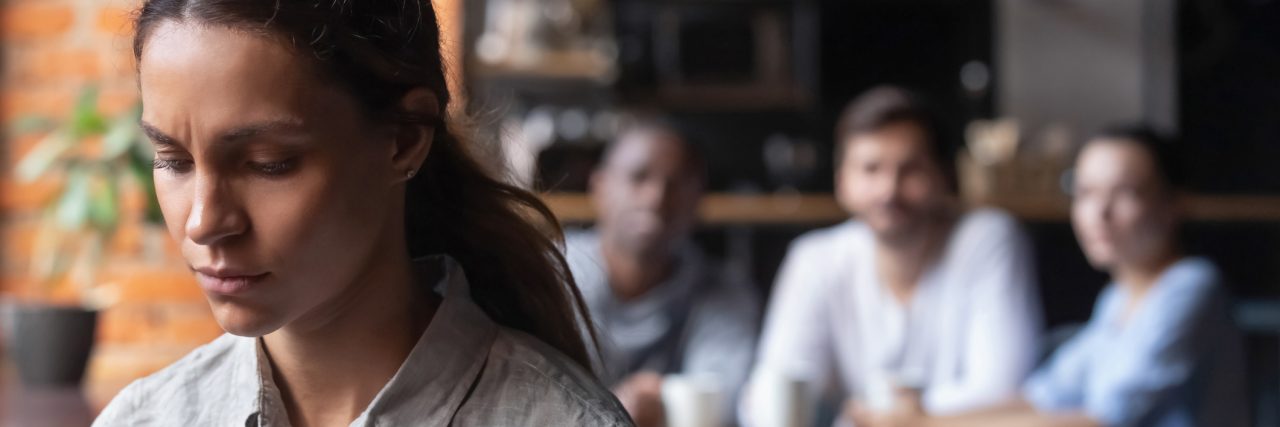 Pulmonary Arterial Hypertension: Feeling Guilty for My Chronic Illness Mixed race woman sitting by herself in a café. She is looking downwards. There is a group of three people at the table behind her.