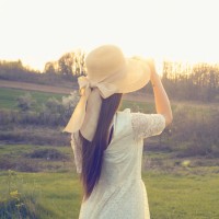 a woman in a white dress and yellow hat outdoors look out at a landscape with field and trees
