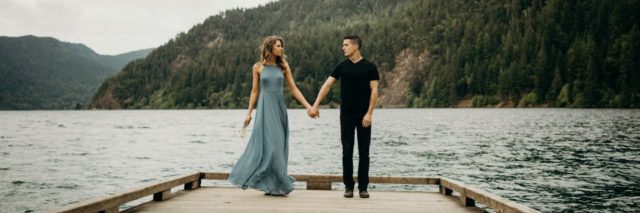 photo of couple holding hands on pier or jetty over lake with mountains in background