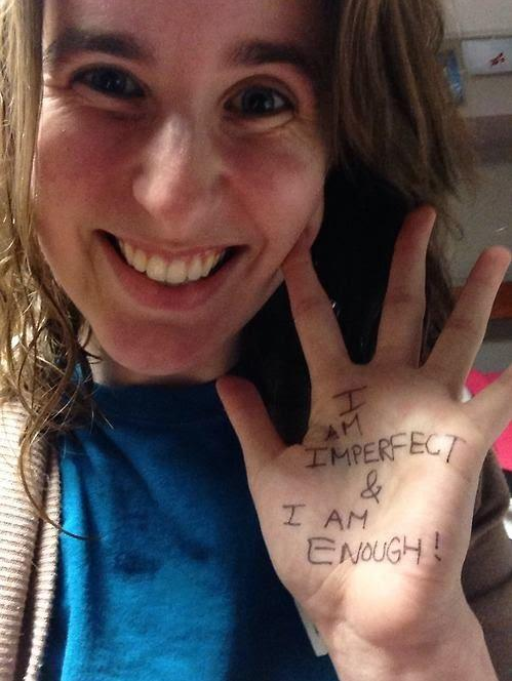Emmie smiling, and holding up the palm of her left hand, on which she wrote in black Sharpie in all caps, “I am imperfect & I am enough!”