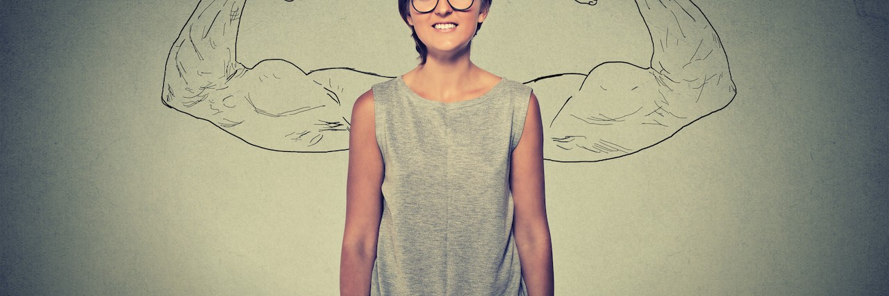 Woman stands in front of a wall with a drawing of two arms flexed, making a muscle behind her