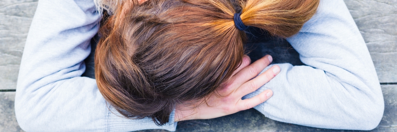Chronic Illness: What Feeling ‘Better’ Looks Like A young woman with her head resting on her hands at a table in the park