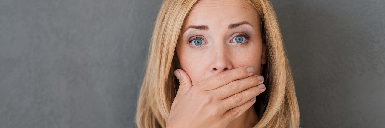 Surprised woman covering mouth with hand and staring at camera while standing against grey background