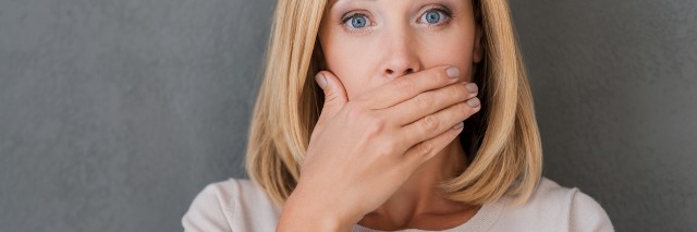 Surprised woman covering mouth with hand and staring at camera while standing against grey background