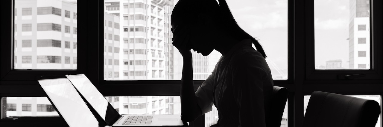 A photographic image of a stressed businesswoman sitting at a desk. The woman is seated in front of a window with a bright light source, rendering her image as a shadowed silhouette with indiscernible features. She is leaning forward with her elbows against the top of the desk and her hands clasped against her face, with her head slightly bent forward.