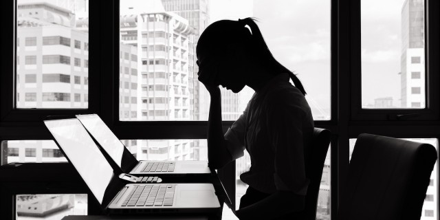 What I Wish I Was Brave Enough to Say at My Lowest Moments A photographic image of a stressed businesswoman sitting at a desk. The woman is seated in front of a window with a bright light source, rendering her image as a shadowed silhouette with indiscernible features. She is leaning forward with her elbows against the top of the desk and her hands clasped against her face, with her head slightly bent forward.