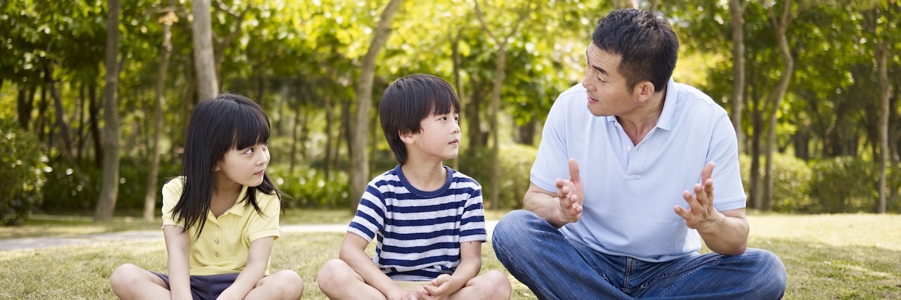 Why We Should Teach Our Kids About Mental Health Father talks to daughter and son as tehy sit outside on grass