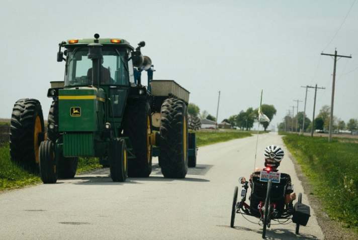 Ray pedals his trike next to a giant tractor. Ray says, “[I rode] 31 miles last Tuesday. Not my usual 100, but ALS has given me the time to absorb my surroundings.”