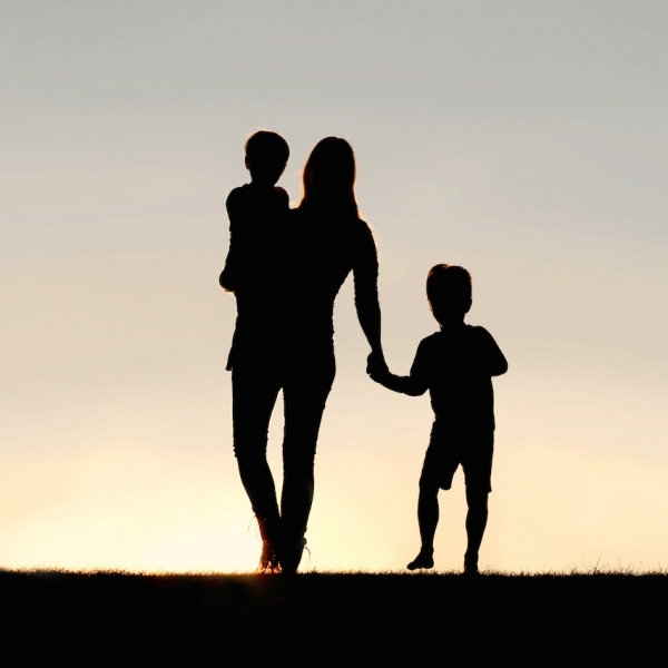 Silhouette of a young mother walking and lovingly holding hands with her happy little child, while holding his baby brother, outside in front of a sunset in the sky.